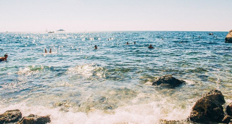 Mar azul con personas nadando cerca de unas rocas, en un día soleado