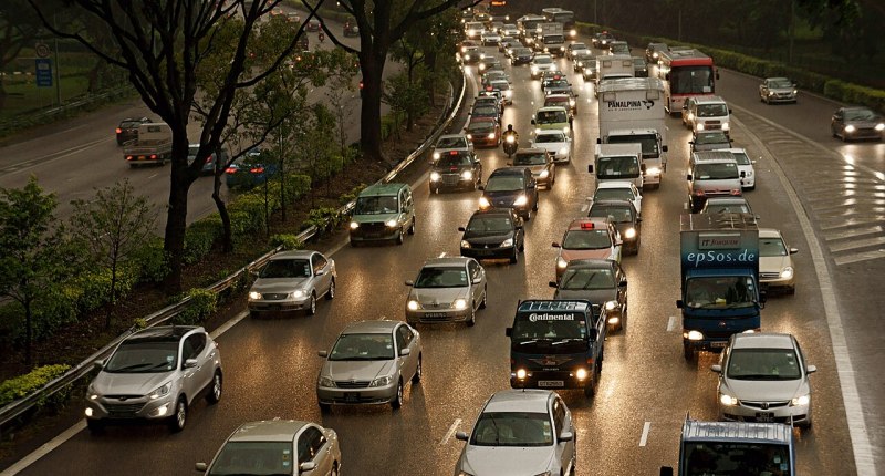 Carros en fila en una vía con lluvia, con luces encendidas y árboles a un costado