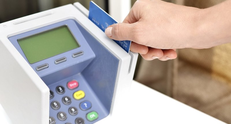 Mano de una persona pasando una tarjeta bancaria azul por un datáfono blanco, en un punto de pago