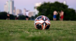 Balón de fútbol naranja, negro y blanco sobre el césped, con edificios borrosos al fondo