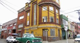 Edificio de esquina amarillo y de ladrillo en un barrio de Bogotá, con casas y locales pequeños a los lados y un carro verde estacionado