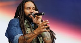 Ky-Mani Marley cantando con micrófono en un escenario de festival, con camisa de mezclilla y chaleco militar