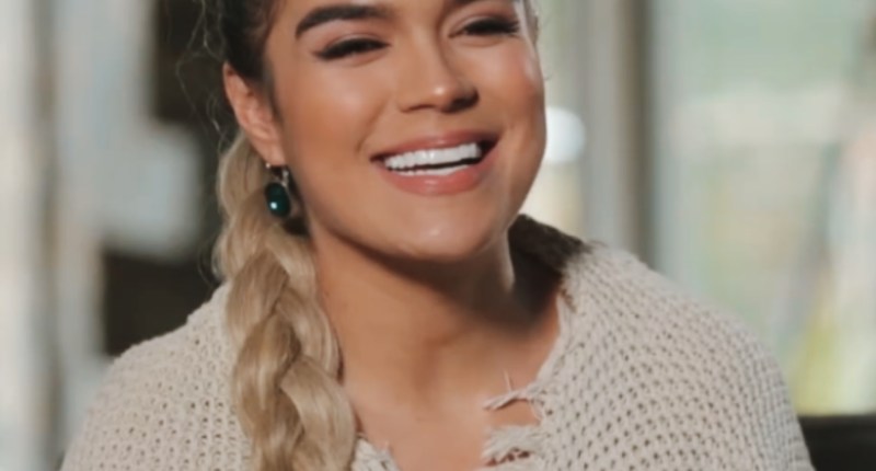 Karol G sonriendo, con una trenza y un suéter de tejido beige, en una entrevista