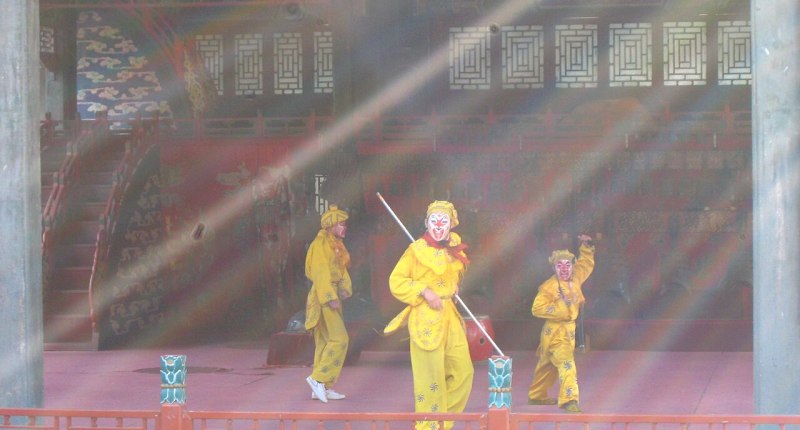 Tres actores de ópera de Pekín con trajes amarillos y máscaras pintadas, en un escenario de madera con decoración roja