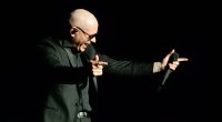 Pitbull cantando en el escenario con un micrófono y gesto con las manos, sobre fondo negro