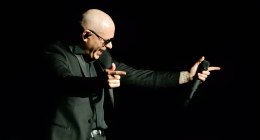 Pitbull cantando en el escenario con un micrófono y gesto con las manos, sobre fondo negro