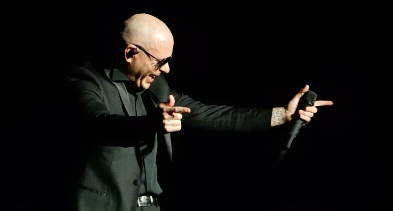 Pitbull cantando en el escenario con un micrófono y gesto con las manos, sobre fondo negro