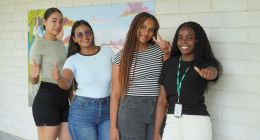 Cuatro jóvenes sonrientes posan frente a un mural en un campus, dos con el pulgar arriba