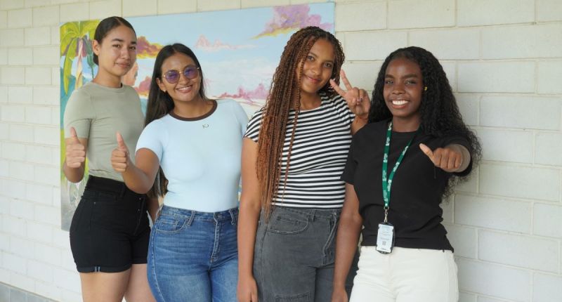 Cuatro jóvenes sonrientes posan frente a un mural en un campus, dos con el pulgar arriba