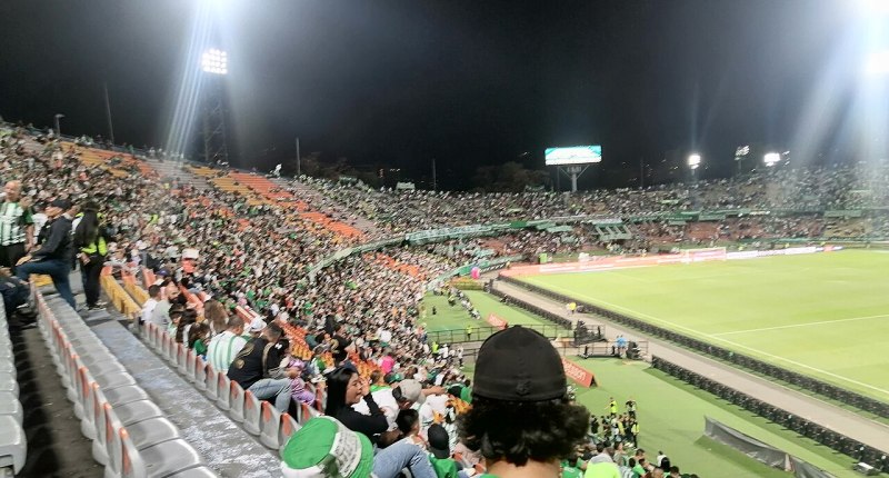 Tribuna sur del Estadio Atanasio Girardot de Medellín de noche, con hinchas de verde y la cancha iluminada