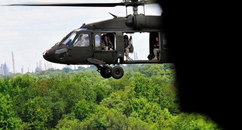 Helicóptero UH-60 Black Hawk del Ejército de EE. UU. en vuelo sobre un bosque, con la puerta abierta y soldados lejanos en el interior