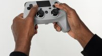 Manos sosteniendo un control de videojuego gris con palancas y botones, sobre fondo blanco