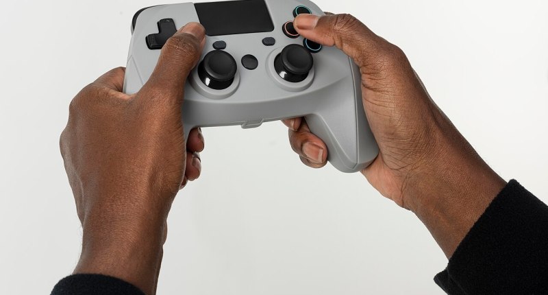 Manos sosteniendo un control de videojuego gris con palancas y botones, sobre fondo blanco