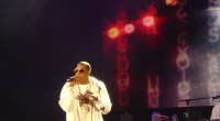 Jay-Z con gafas oscuras y chaqueta blanca, cantando con micrófono en un escenario oscuro con luces rojas