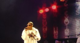 Jay-Z con gafas oscuras y chaqueta blanca, cantando con micrófono en un escenario oscuro con luces rojas