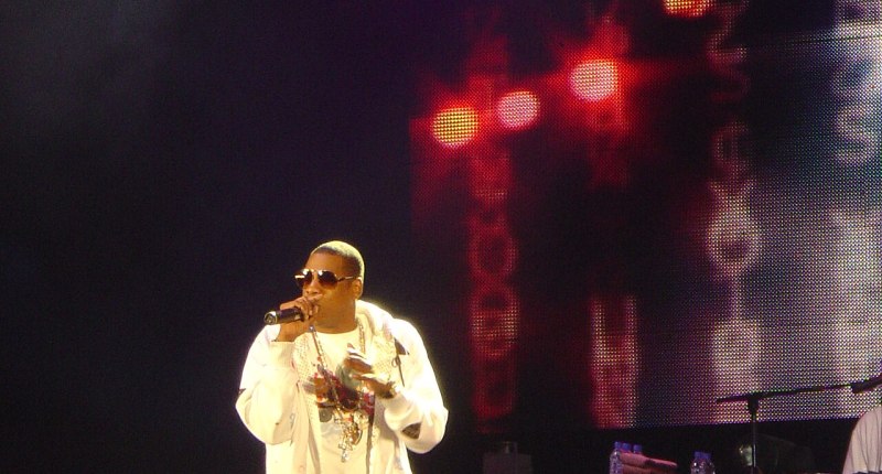 Jay-Z con gafas oscuras y chaqueta blanca, cantando con micrófono en un escenario oscuro con luces rojas