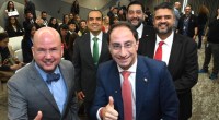 Cinco hombres con traje sonríen en una sala con asistentes detrás; dos hacen pulgar arriba