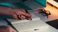 Manos escribiendo en una libreta junto a un portátil y un celular sobre un escritorio
