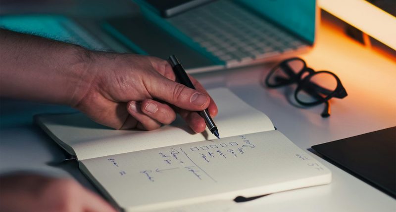 Manos escribiendo en una libreta junto a un portátil y un celular sobre un escritorio