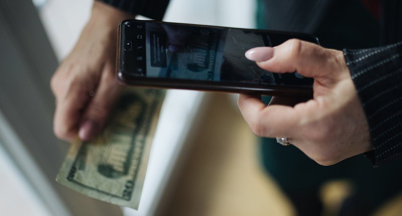 Manos de una persona fotografiando un billete de dólar con su celular, en una escena de pago