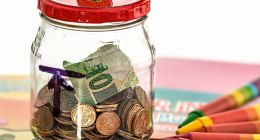 Frasco de vidrio con monedas y un billete de papel dentro, con la tapa roja, sobre una mesa con crayones de colores