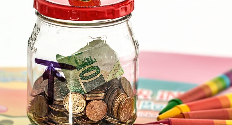 Frasco de vidrio con monedas y un billete de papel dentro, con la tapa roja, sobre una mesa con crayones de colores