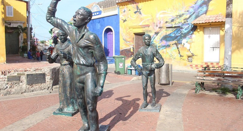 Estatuas de bronce en la Plaza de la Trinidad de Getsemaní, Cartagena, con un mural de pájaro en la fachada amarilla