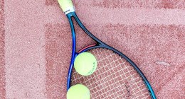 Raqueta de tenis azul y dos pelotas amarillas sobre una cancha de arcilla roja