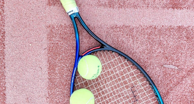 Raqueta de tenis azul y dos pelotas amarillas sobre una cancha de arcilla roja