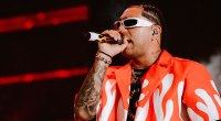 Ryan Castro cantando con micrófono en la mano, gafas blancas y camisa blanca con llamas rojas, frente a luces rojas de escenario