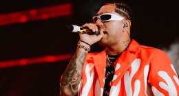 Ryan Castro cantando con micrófono en la mano, gafas blancas y camisa blanca con llamas rojas, frente a luces rojas de escenario