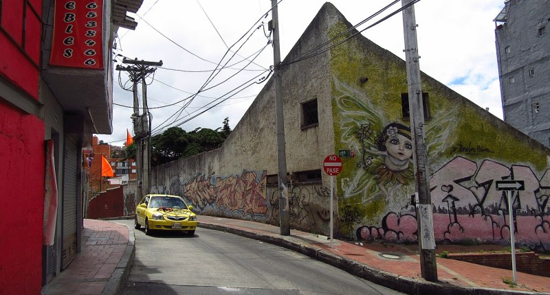 Taxi amarillo en una calle empinada de Bogotá, con muros grafiteados y un mural al fondo