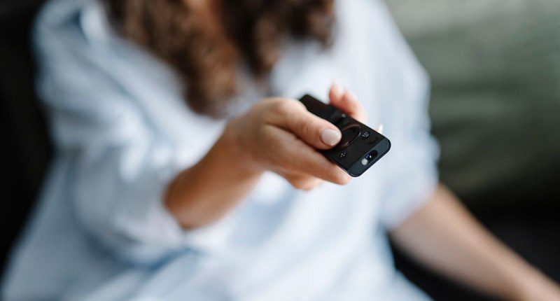 Persona sentada en un sofá apuntando con un control remoto negro hacia el televisor, en una casa