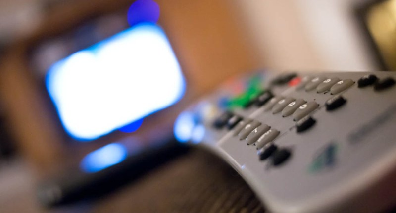 Control remoto gris en primer plano, con un televisor encendido desenfocado al fondo