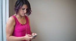 Mujer de pelo morado y blusa fucsia escribe en su celular junto a una pared clara